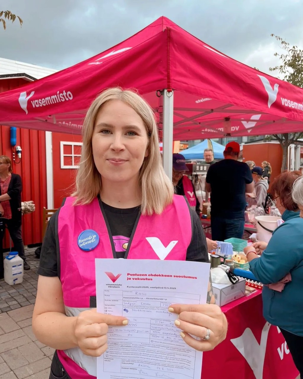 Raision Vasemmistoliiton kuntavaaliehdokas Emma Lindqvist pitää kädessään kuntavaalien ehdokassuostumuslomaketta. Taustalla näkyy Vasemmistoliiton teltta ja Killin Markkinat.